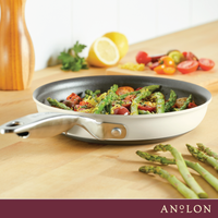 A pan filled with asparagus, tomatoes, and seasoning sits on a wooden countertop. Fresh asparagus spears and halved tomatoes are nearby. A cut lemon and parsley are in the background. The brand Anolon is visible on the bottom border.