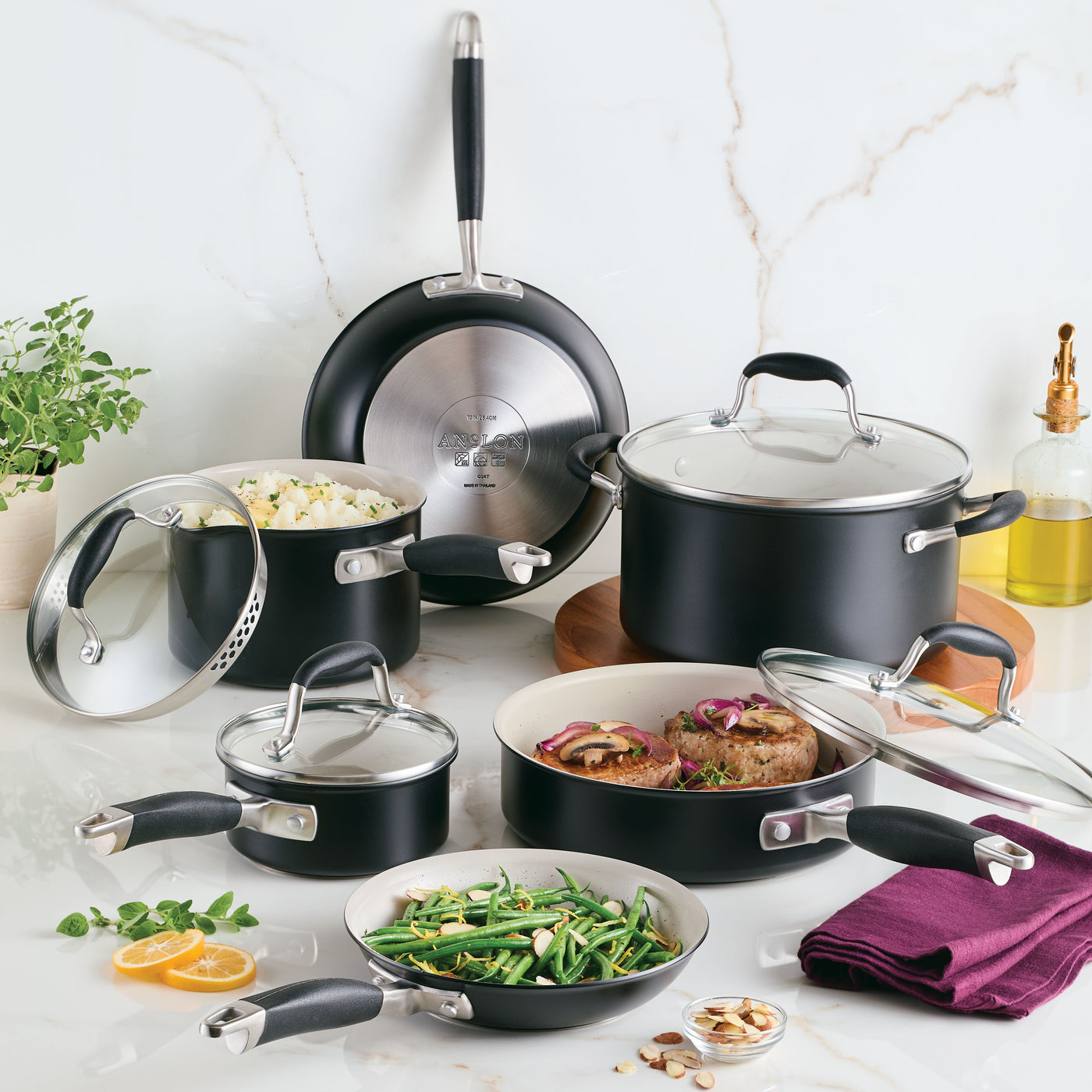 A 10-Piece Ceramic Nonstick Cookware Set by Advanced Ceramic is showcased on a marble counter. The black and stainless steel pots and pans hold assorted cooked dishes, such as vegetables and meat. In the background, theres a small plant, lemon slices, and a bottle of oil.