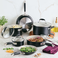 A 10-Piece Ceramic Nonstick Cookware Set by Advanced Ceramic is showcased on a marble counter. The black and stainless steel pots and pans hold assorted cooked dishes, such as vegetables and meat. In the background, theres a small plant, lemon slices, and a bottle of oil.