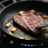 Close-up of a steak sizzling in the Anolon X 10-Inch Hybrid Nonstick Frying Pan with garlic cloves and rosemary. Tongs hold the steak as it browns, while the stovetop burner is visible beneath, highlighting its induction-suitable design.