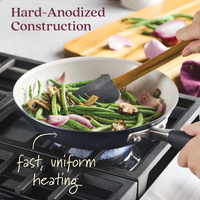 A hand uses a spatula to stir green beans, onions, and mushrooms in a small pan on a stovetop. The Advanced Ceramic 2-Piece Frying Pan Set features hard-anodized construction and fast, uniform heating, ensuring quality performance.