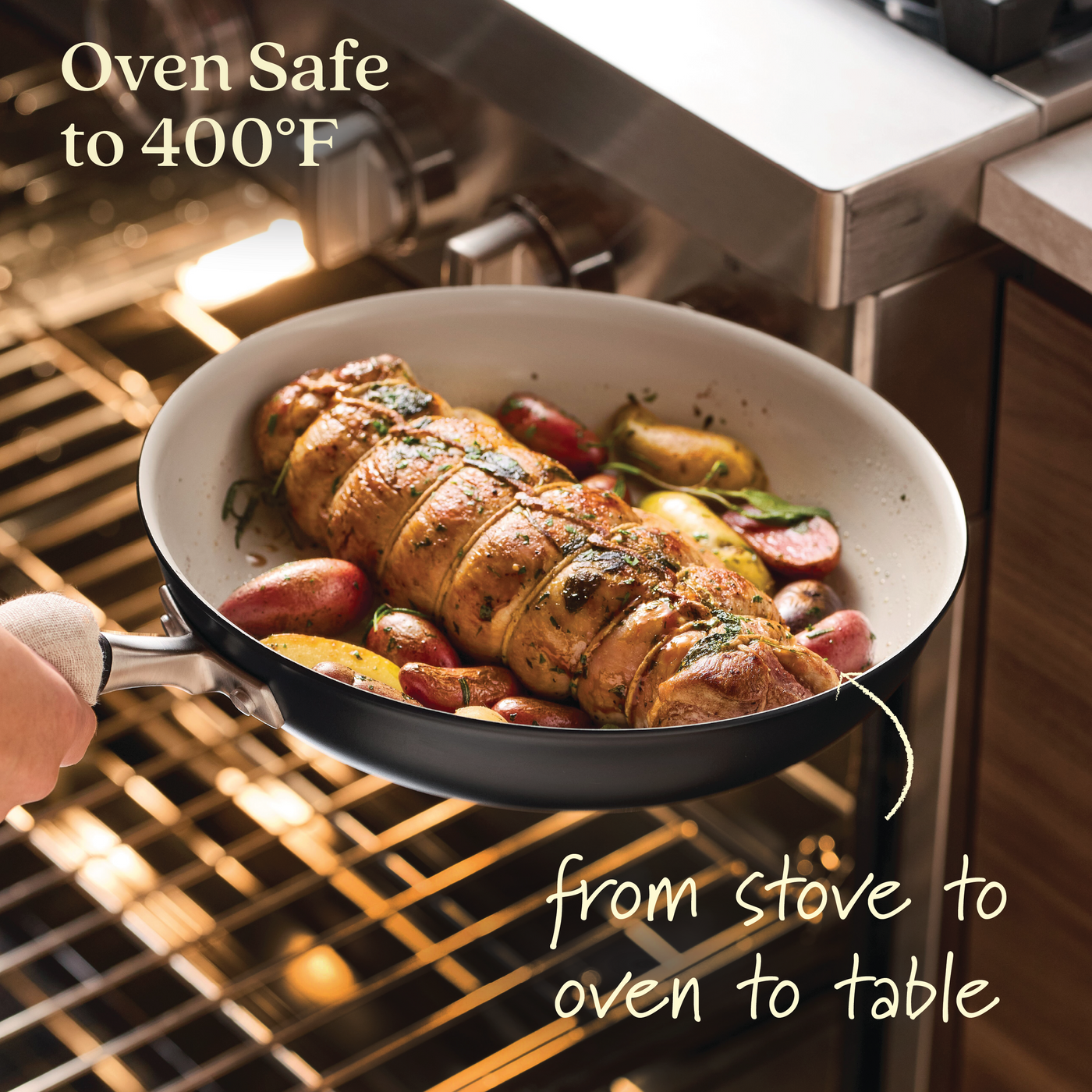 A hand places an Advanced Ceramic 12-inch Nonstick Frying Pan with a seasoned meat roast and vegetables into an oven. The cookware is oven-safe up to 400°F, seamlessly transitioning from stove to table. It features a sleek black exterior with a white interior.