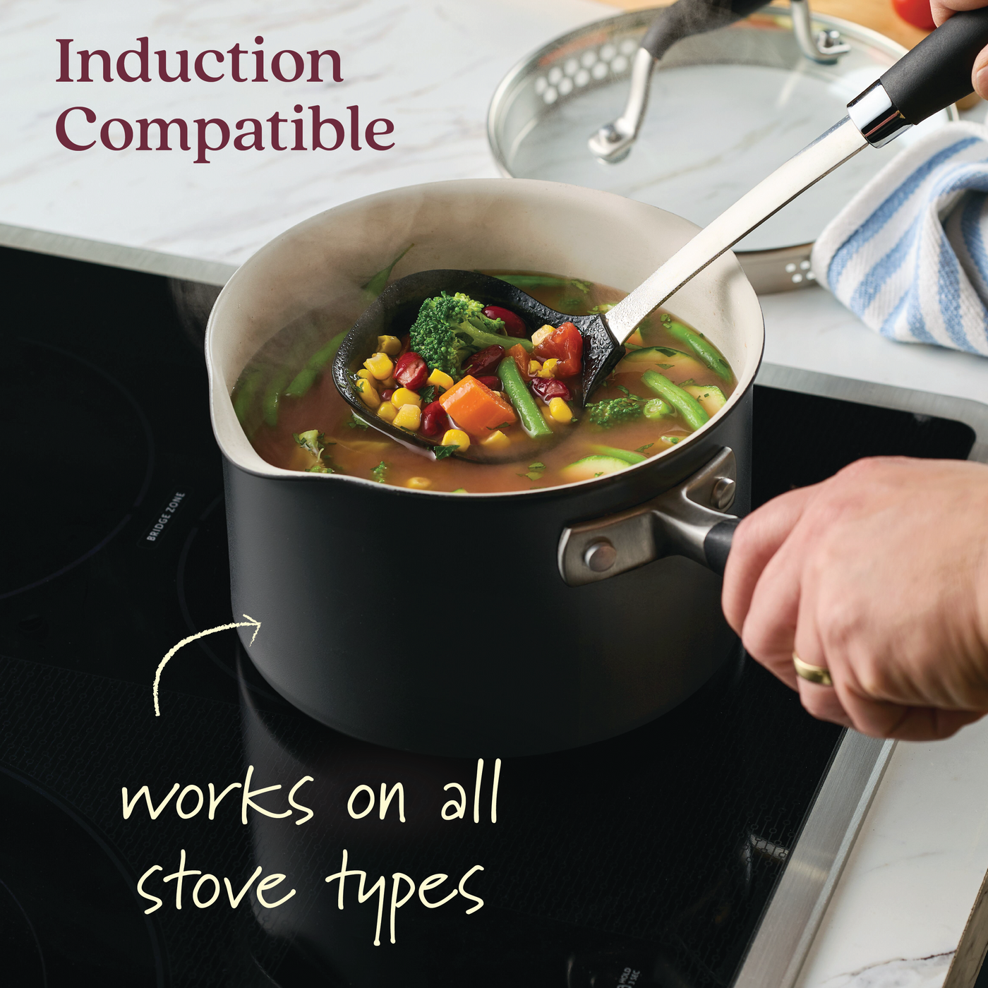 Someone stirs vegetable soup in a black saucepan from the Advanced Ceramic 10-Piece Nonstick Cookware Set on an induction stove. The image reads, Induction Compatible and Works on all stove types, with a striped cloth and kitchen utensils in the background.