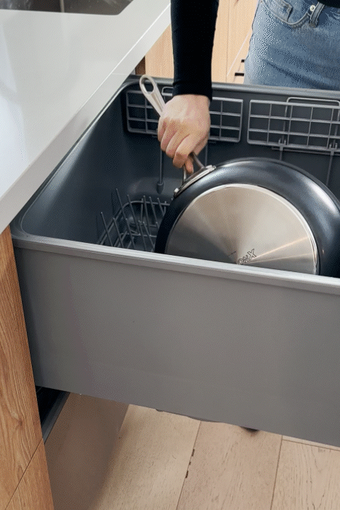 A short, looping GIF demonstrating the convenience of Anolon X cookware. A person is shown placing a large frying pan with a stainless steel base into the bottom rack of a modern dishwasher, highlighting that the collection is dishwasher safe for easy cleanup.