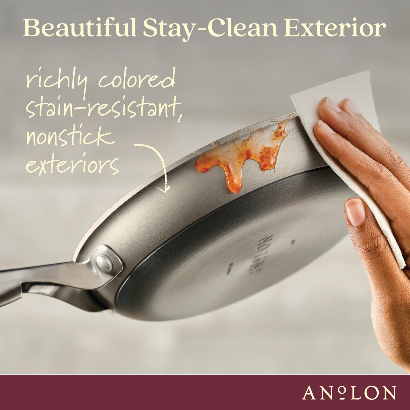 A hand wipes food off a frying pan with a cloth, showcasing the pans stain-resistant and nonstick exterior. Text on the image reads Beautiful Stay-Clean Exterior and highlights its richly colored, easy-to-clean surface.