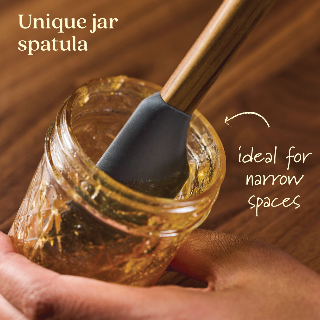 A hand holds a glass jar with a brown substance, using a spatula from the Tools and Gadgets 3-Piece Spatula Set, featuring a teak handle and dark silicone head. The image text reads, "Perfect for narrow spaces with this unique jar spatula.