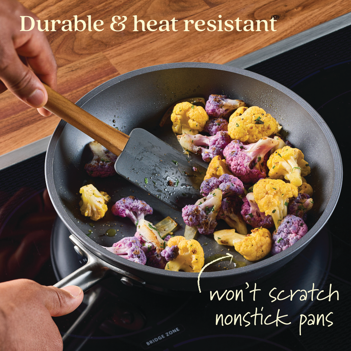 A hand using a spatula from the Tools and Gadgets 3-Piece Spatula Set gently stirs sautéed purple and yellow cauliflower in a nonstick pan. Text reads, Durable & heat resistant and won't scratch nonstick pans. The pan is on an induction stove with a wooden countertop.