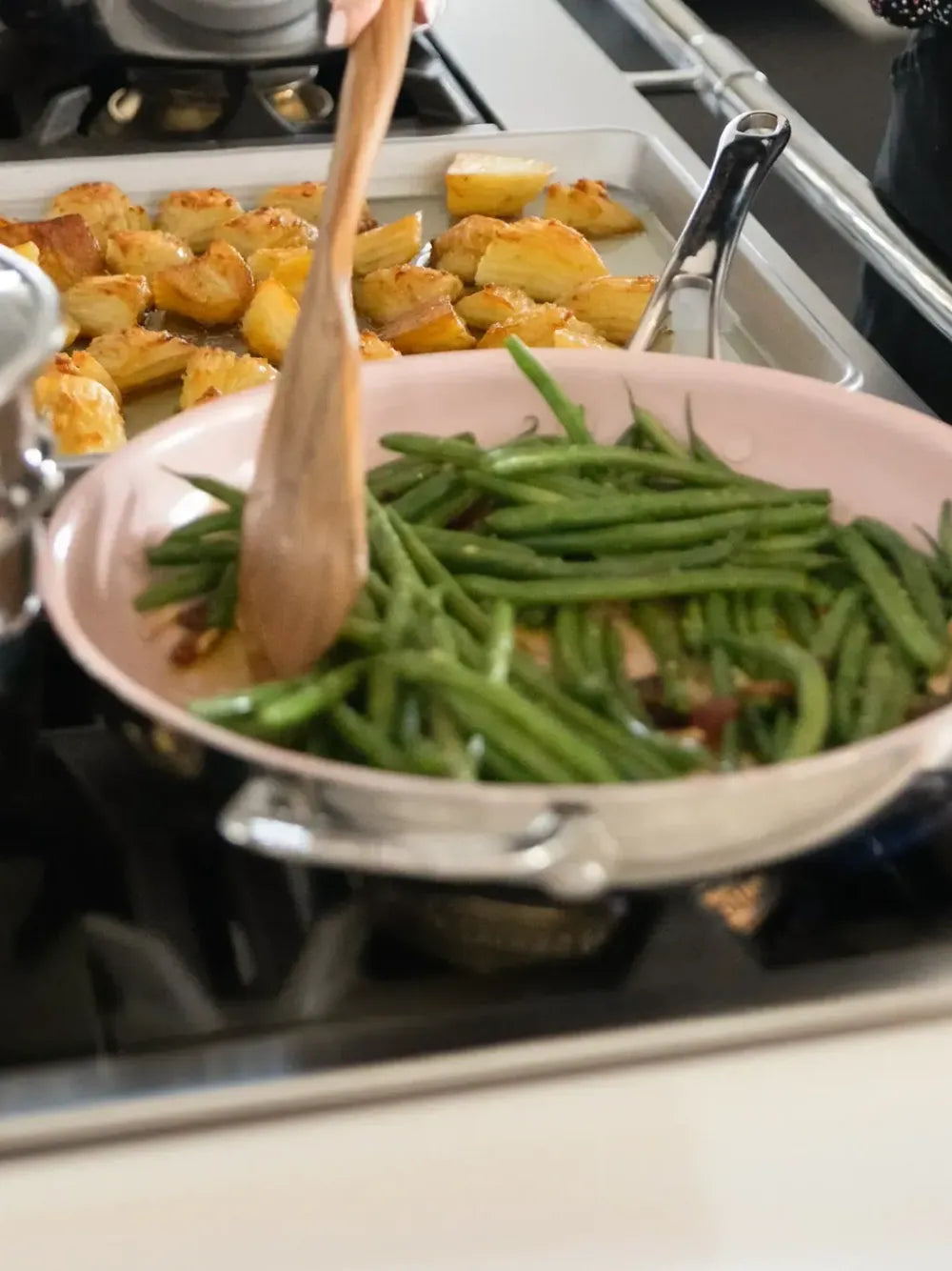 A pink ceramic pan on a stovetop being stirred with a wooden spoon, containing green beans, with roasted potatoes in a baking pan in the background.