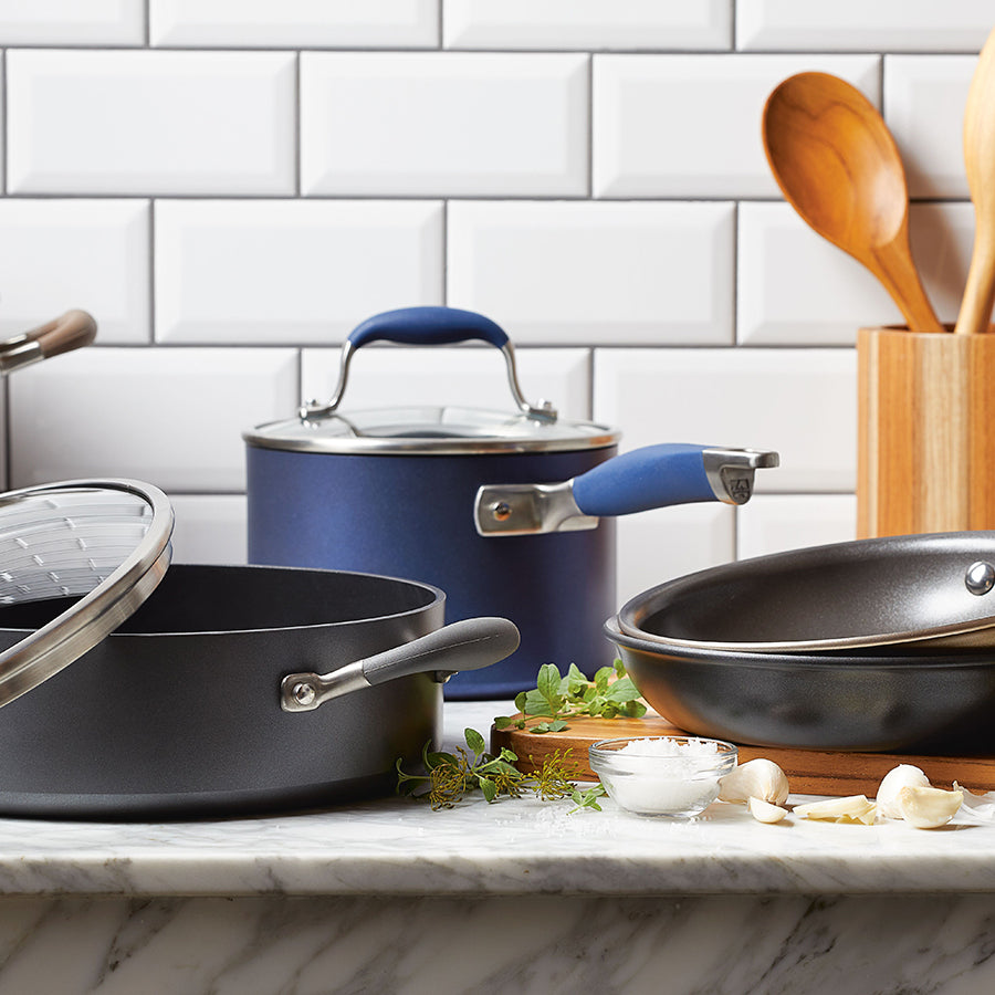 A collection of Anolon Advanced Home Cookware is shown on a wooden cutting board against a white tiled backsplash. The set features dark gray nonstick surfaces with coordinating navy blue SureGrip handles and includes a large stir-fry pan with a glass lid, a smaller frying pan, and a stockpot. Fresh asparagus, garlic, and a small bowl of salt are arranged around the pans, with wooden utensils stored in a block in the background.