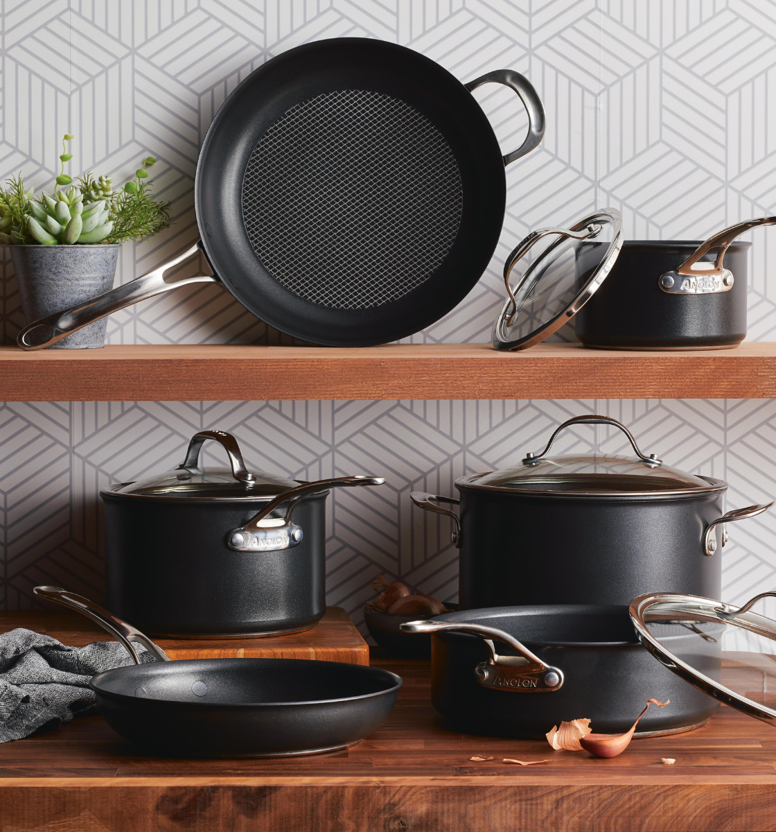 A stylish kitchen display features various black cooking pots and pans with stainless steel handles arranged on wooden shelves. A small green succulent adds decoration, and the background showcases a geometric-patterned wall.