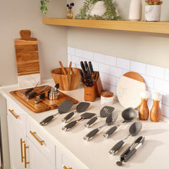 A kitchen countertop showcases the Exclusive Everything Set: 75 Pieces, with an array of cooking utensils like spatulas and tongs neatly arranged. A knife block sits beside a wooden decorative board, accompanied by bowls, salt and pepper shakers, plants, and advanced SearTech™ technology to create a warm and organized atmosphere.