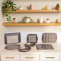 A kitchen counter showcases an assortment of baking trays and pans from The Everything Set: 75 Pieces by Exclusive. Open shelves above hold potted plants and decorative vases, while white cabinets and a tiled backsplash, enhanced with SearTech™ technology, create a clean, organized look.