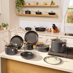 A kitchen display presents the 75-piece Everything Set from Exclusive, featuring a complete selection of cookware such as pots, pans, and lids elegantly arranged on the countertop and stove. The Anolon X line adds to the ambiance against a tiled backsplash with potted plants as sunlight streams through the window.