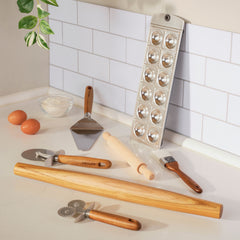 The Exclusive Everything Set: 75 Pieces elegantly adorns the countertop, showcasing baking tools such as a ravioli mold, rolling pin, pastry brush, pizza cutter, dough scraper, and fluted pastry wheel. Nearby sit two eggs and a small bowl of flour. Overhead, a plant adds to this culinary display.