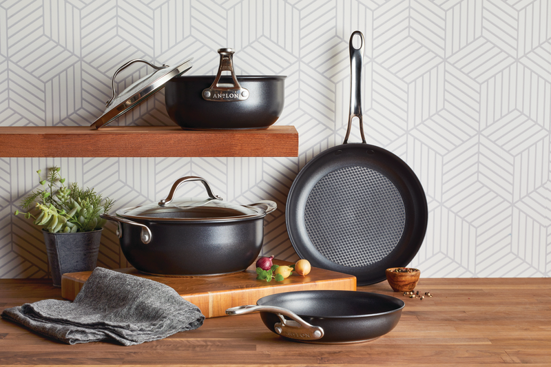 A collection of Anolon X Cookware featuring dark gray exteriors and distinctive stainless steel mesh grids on the nonstick cooking surfaces. The set includes a lidded saucepan on a wooden shelf, a larger lidded pot, and two frying pans—one standing upright to show the grid pattern and another resting on a wooden countertop alongside a gray kitchen towel, fresh herbs, and small bowls of ingredients.