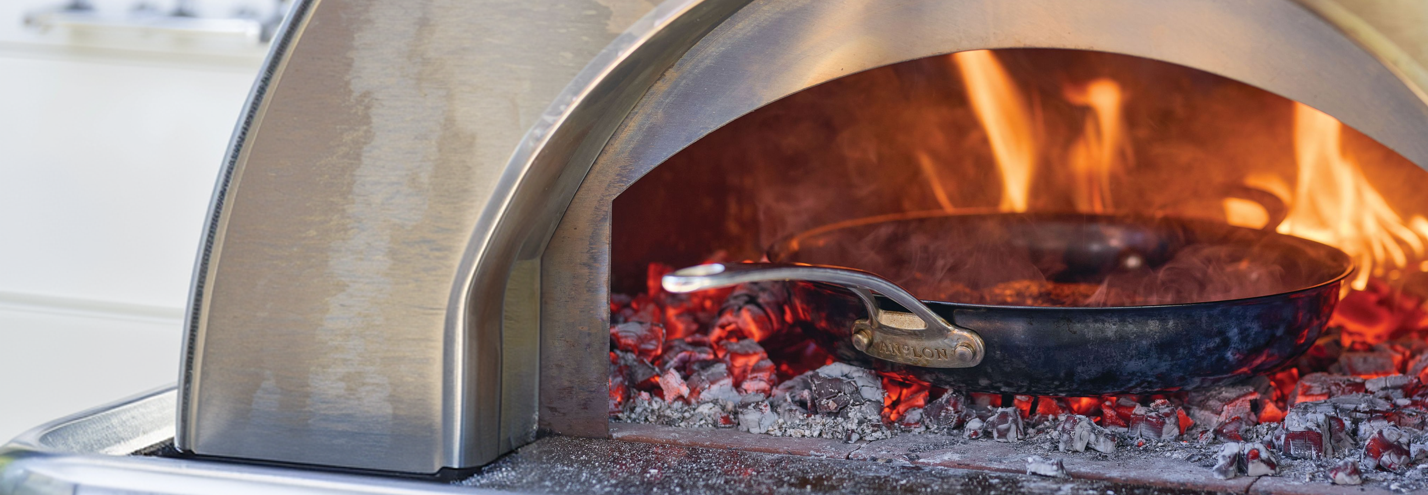 An Anolon carbon steel frying pan sits inside a pizza oven, surrounded by glowing red embers and orange flames.