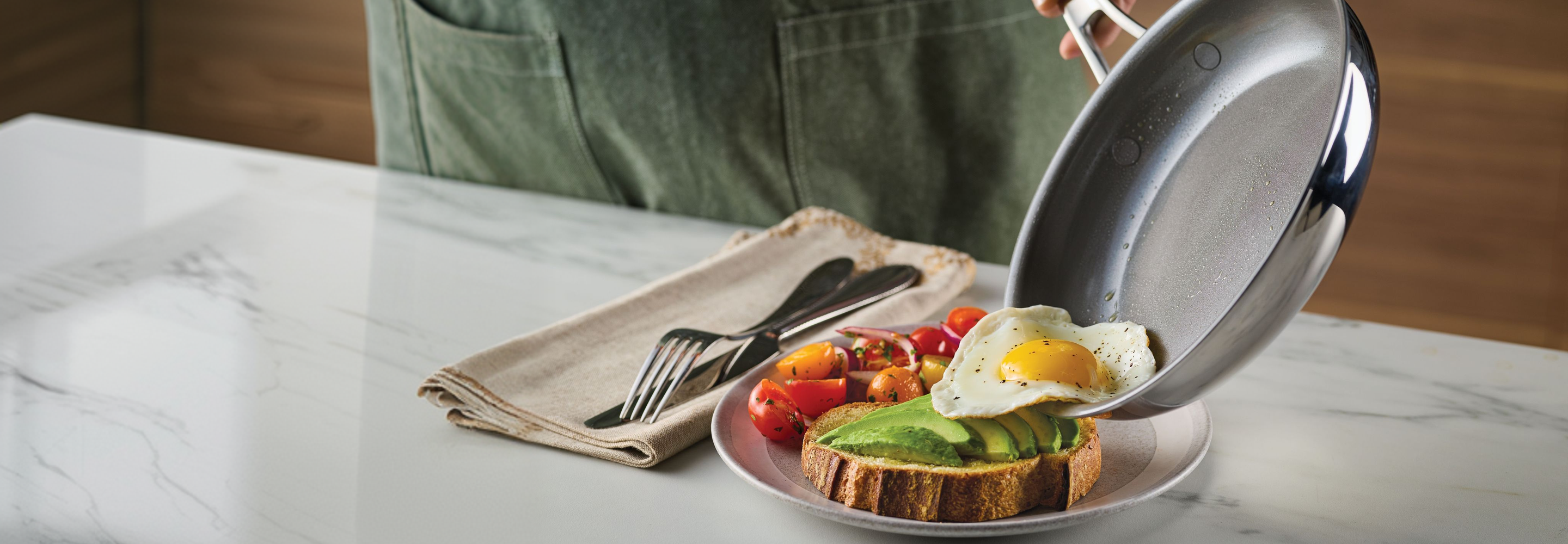A person sliding a fried egg from an Anolon EverLast Clad Ceramic frying pan onto avocado toast with roasted tomatoes.