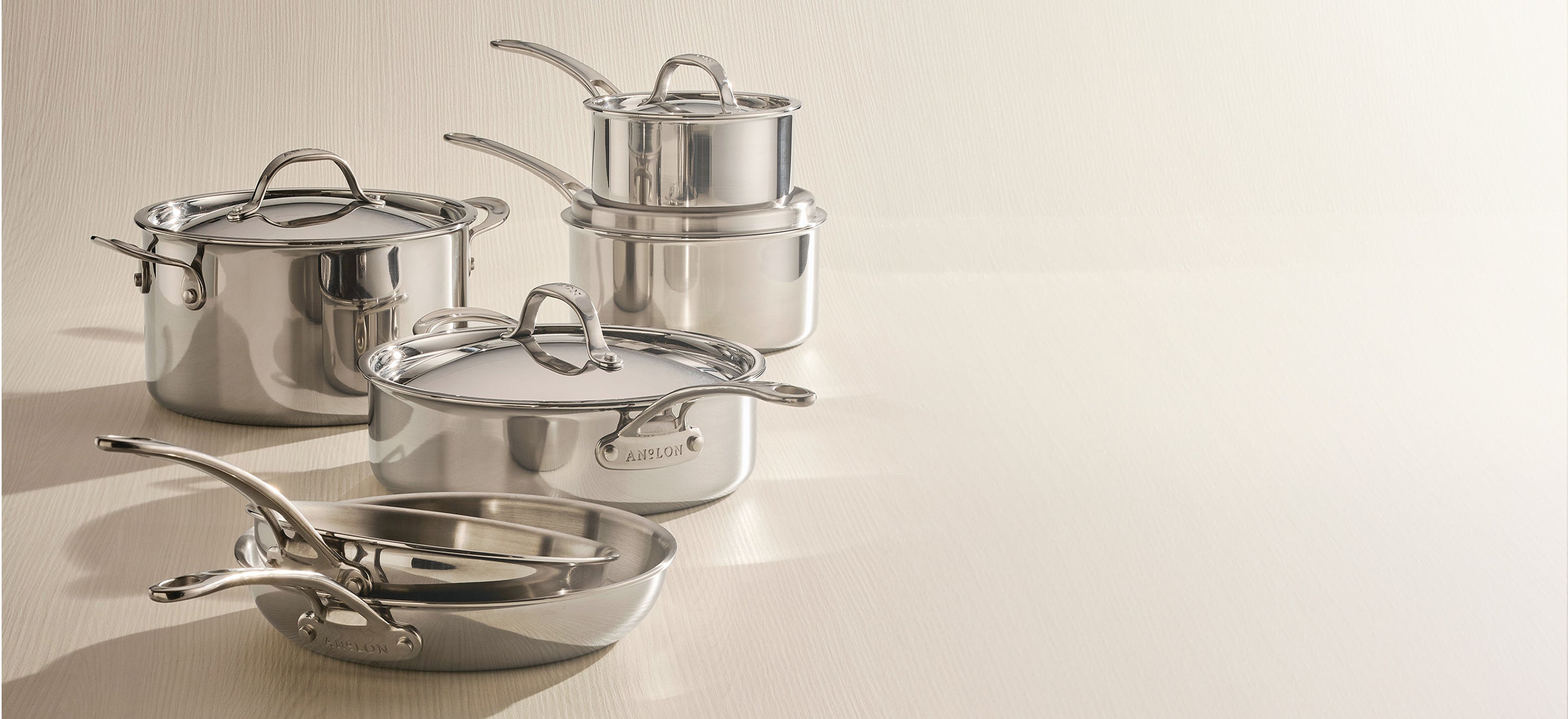 A studio photograph of the Anolon EverLast Clad Stainless Steel 10-piece cookware set. The polished tri-ply pieces, including a large stockpot, a sauté pan, and nested saucepans with lids, are elegantly arranged on a light-colored surface against a neutral, minimalist background, highlighting their professional luster and modern design.