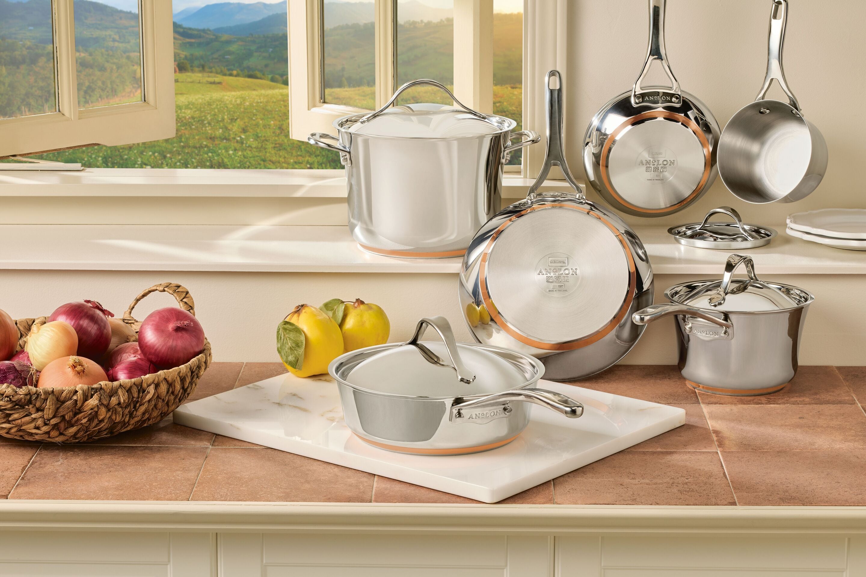A lifestyle photograph of a 10-piece Anolon Nouvelle Copper Stainless Steel cookware set arranged on a kitchen counter in front of a window. The set includes various sized pots and pans with a polished mirror finish, stainless steel lids, and the signature copper ring visible at the base. A basket of onions and a few lemons sit nearby on the marble countertop, creating a bright, culinary atmosphere.
