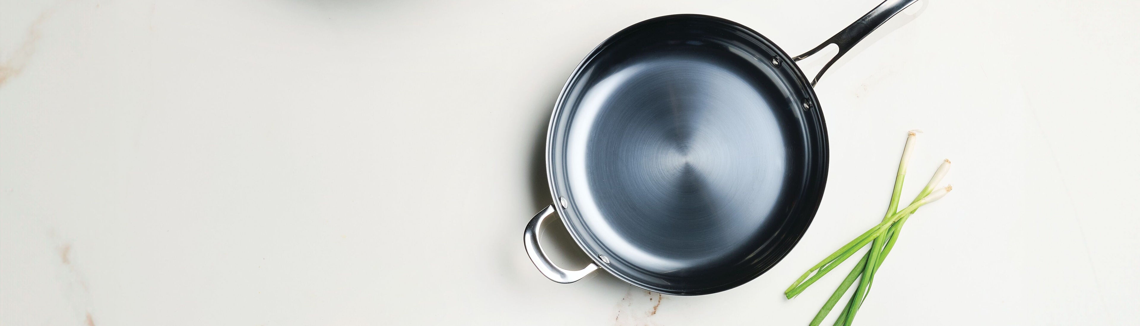 A top-down studio photograph of an Anolon EverLast Carbon Steel 12-inch frying pan. The pan features a dark, pre-seasoned interior and a polished stainless steel handle with a matching helper handle. It is presented on a white marble surface next to a bundle of fresh green scallions, highlighting its sleek profile and professional construction.