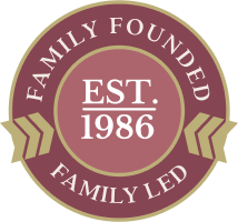 A circular, burgundy-colored badge with a scalloped border and gold ribbon accents on the sides. The center features the text "EST. 1986" in white, surrounded by the words "FAMILY FOUNDED" and "FAMILY LED," highlighting the heritage of the Anolon brand.