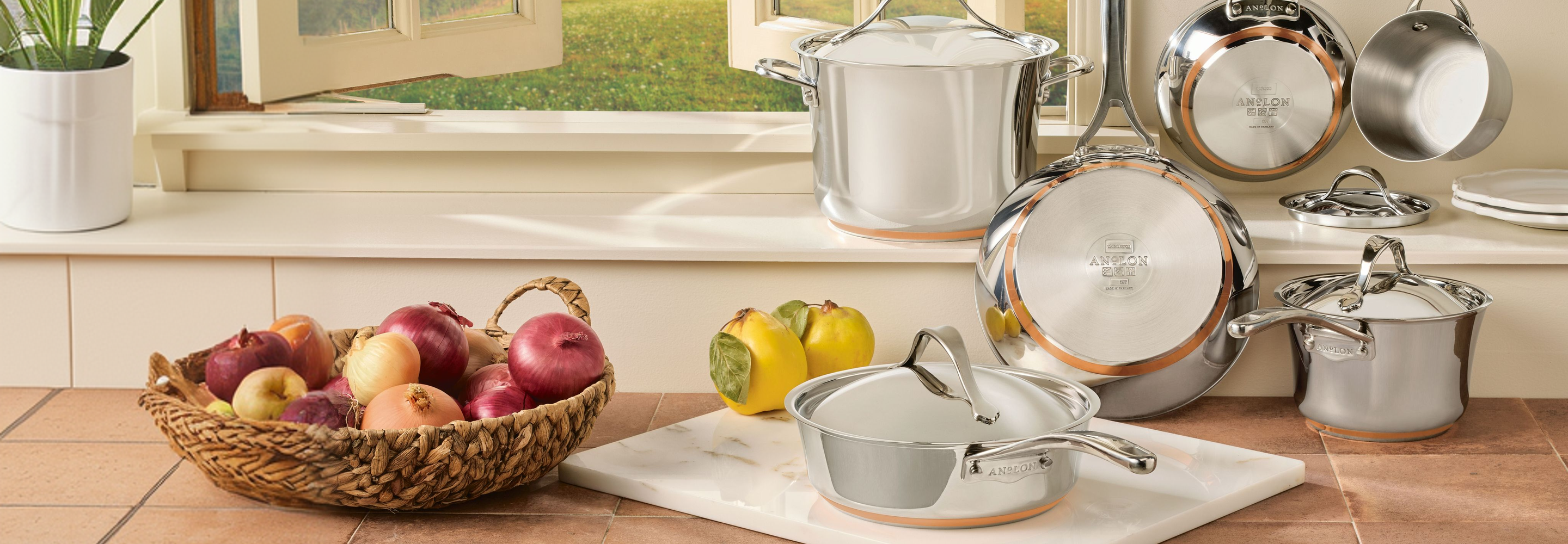 Anolon Nouvelle Copper Stainless Steel cookware set, including stockpots and saucepans with visible copper bases, arranged on a kitchen counter next to a basket of onions.