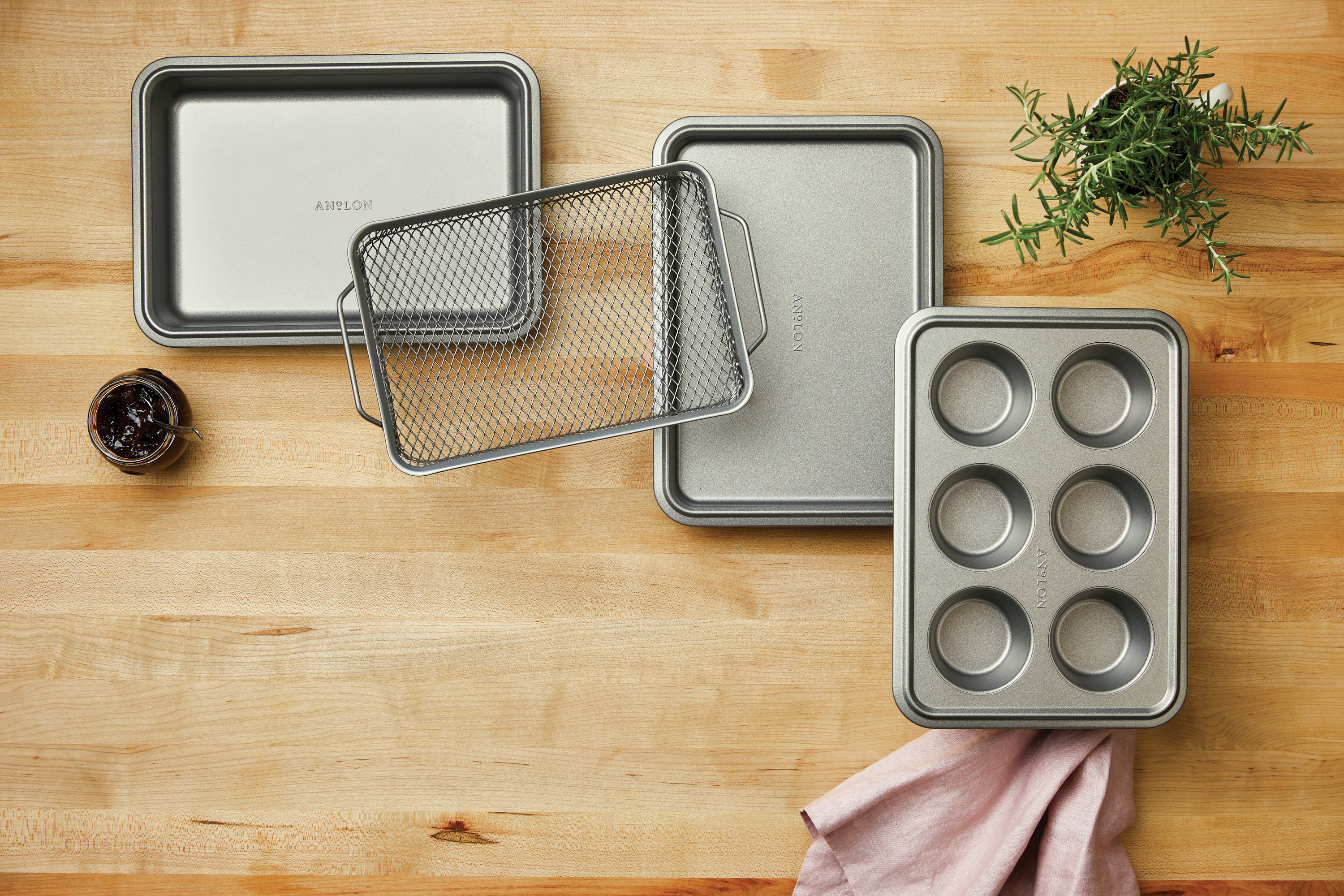 A top-down lifestyle photograph of the  4-piece toaster oven bakeware set arranged on a wooden countertop. The set includes a small cookie sheet, a cake pan, a cooling rack, and a 6-cup muffin tin, all in a dark gray nonstick finish. The scene is accented with a sprig of fresh rosemary and a small jar of jam.