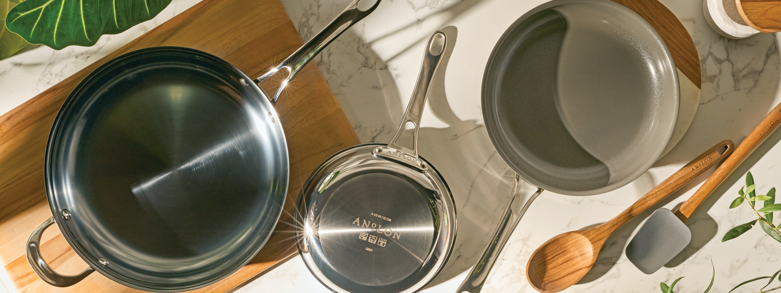 A high-angle, aerial lifestyle photograph of three distinct Anolon EverLast pans arranged on a white marble surface. The collection includes an EverLast Carbon Steel frying pan, a smaller EverLast Clad Stainless Steel saucepan showing the branded base, and an EverLast Clad Ceramic pan with a smooth gray interior. The scene is completed with a wooden cutting board, a wooden spoon, and a gray silicone spatula.