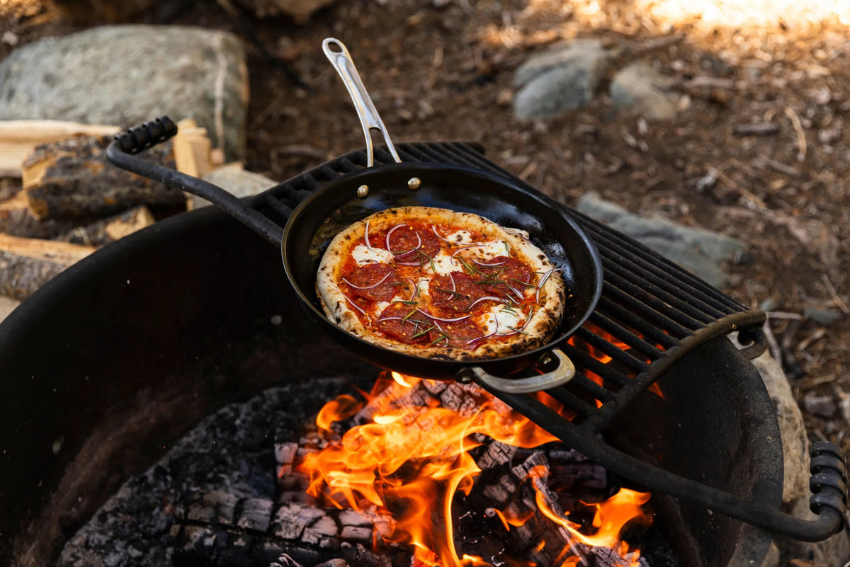 A small black skillet with a pizza cooks over an open campfire on a grill stand outdoors.