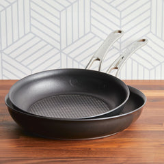 The Anolon X 8.25-Inch and 10-Inch Hybrid Nonstick Frying Pan Set, featuring ultra-durable metal handles and a grid pattern with SearTech™ technology, is stacked on a wooden countertop. The stylish kitchen scene has a light gray and white geometric background.