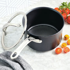 An AnolonX 3-Quart Hybrid Nonstick Saucepan with Lid, featuring ultra-durable nonstick and SearTech™ technology, rests on a marble countertop. Nearby are a few whole and sliced cherry tomatoes in red and yellow, complemented by a gray kitchen towel placed beside it.