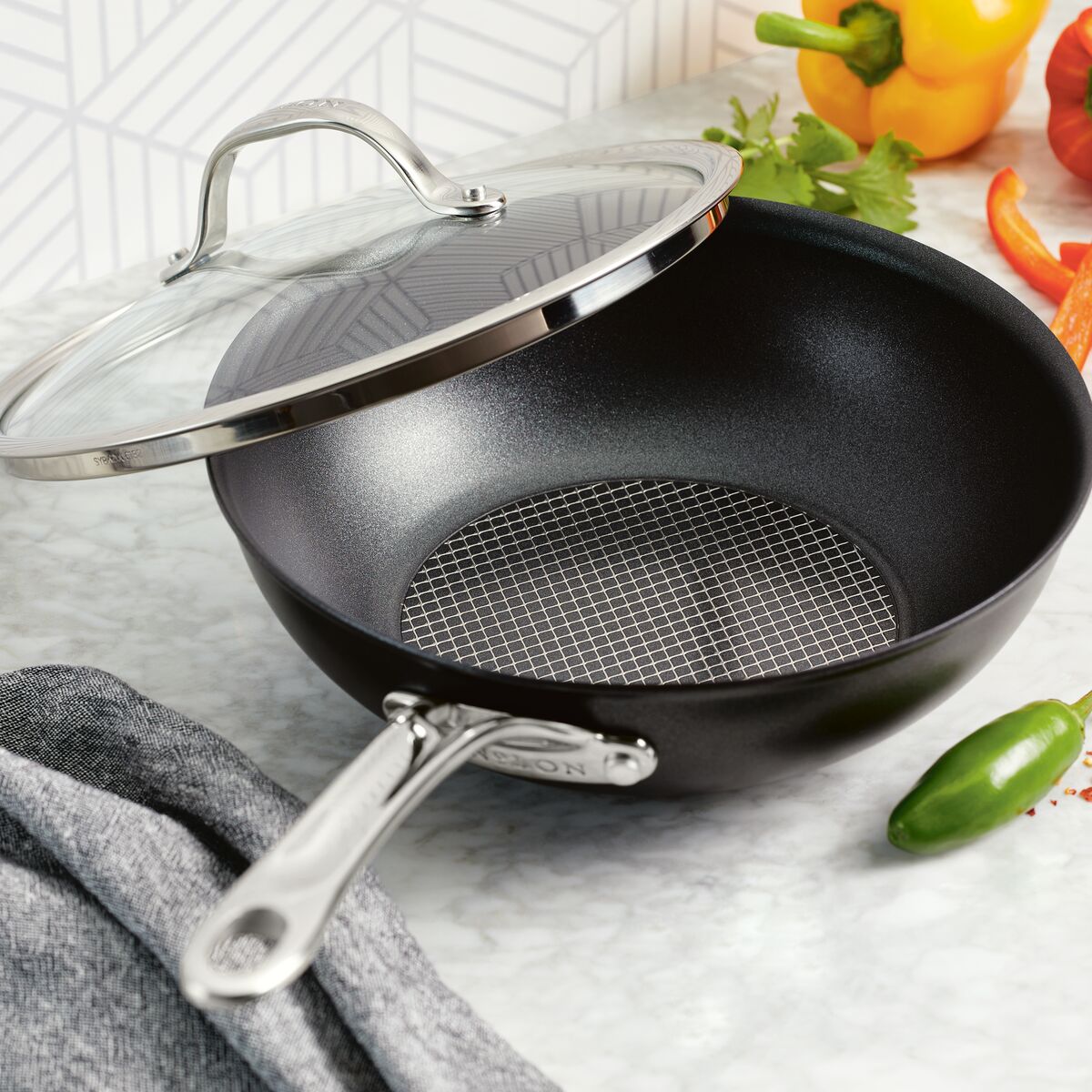 Anolon X 10-Inch Hybrid Nonstick Wok with Lid, a sleek black design on a marble countertop, is surrounded by vibrant veggies—yellow and red bell peppers, jalapeño, cilantro, and sliced carrot. Its stainless steel base suits culinary enthusiasts. Nearby lies a draped gray cloth.