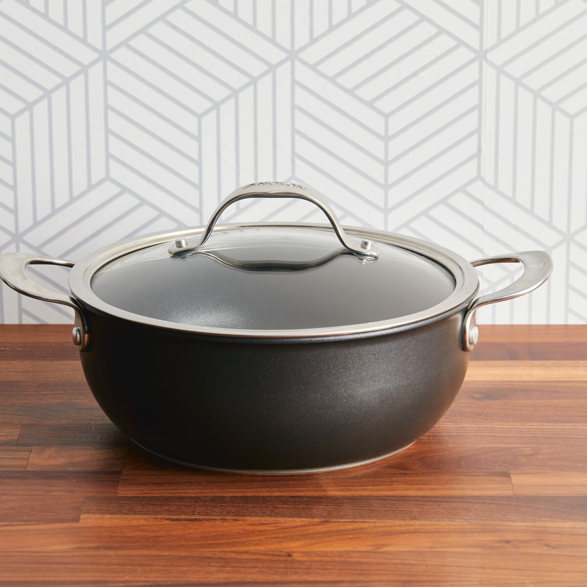 Anolon Xs 4-Quart Hybrid Nonstick Casserole Pan With Lid in black, featuring metal handles and a glass lid, rests on a wooden surface. Its stainless steel base is highlighted by the geometric grey and white patterned wallpaper in the background.