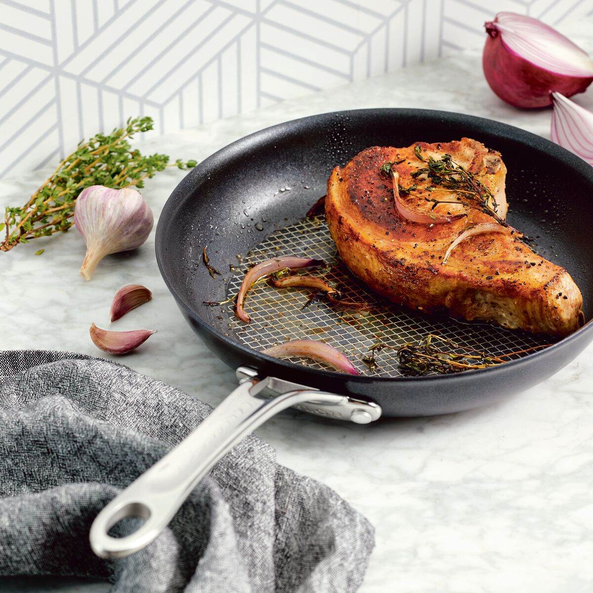 A seasoned pork chop with herbs and onions sizzles in an Anolon X 10-Inch Hybrid Nonstick Frying Pan, surrounded by garlic cloves, thyme sprigs, and a halved red onion on a marble countertop. A gray cloth is partially visible in the bottom corner.