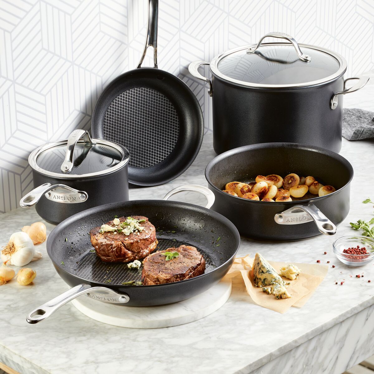 On a marble countertop, the Anolon X 7-Piece Hybrid Nonstick Cookware Set includes a frying pan with steaks, a saucepan, a sauté pan with roasted potatoes, and a stockpot. Fresh herbs, garlic, and blue cheese are scattered around. A geometric patterned wall adds visual interest in the background.