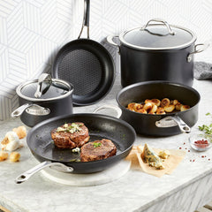 A set of AnolonX 7-Piece Hybrid Nonstick Cookware sits proudly on a marble countertop, displaying its capabilities as two steaks sizzle with even browning thanks to SearTech™ technology, amid garlic and herbs. Meanwhile, cooked potatoes rest in another pan, with a stockpot and saucepan nearby against a geometric wall.