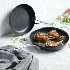 Two sleek frying pans from Anolon Xs 10-Inch and 12-Inch Hybrid Nonstick Set hold rosemary-garnished lamb chops on a marble surface beside a gray cloth, garlic, and herbs. The stylish pans enhance the geometric white tile background.