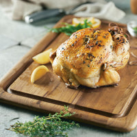 A whole roasted chicken garnished with herbs is served on the Pantryware 20 x 14-Inch Reversible Teak Cutting Board, accompanied by lemon wedges and fresh thyme.