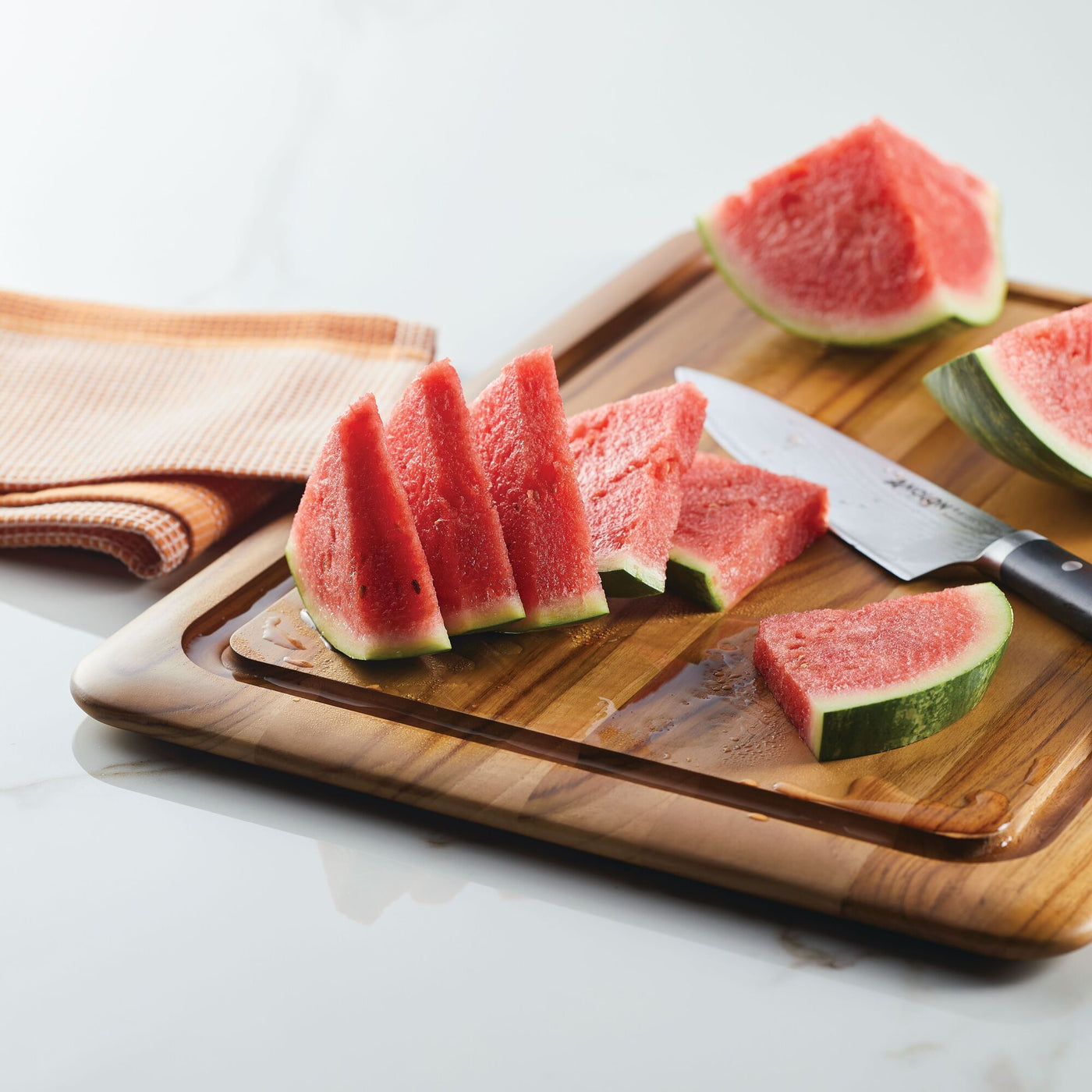 Slices of fresh watermelon sit on the Pantryware 20 x 14-Inch Reversible Teak Cutting Board with a knife. An orange and white towel is folded on the white countertop nearby.