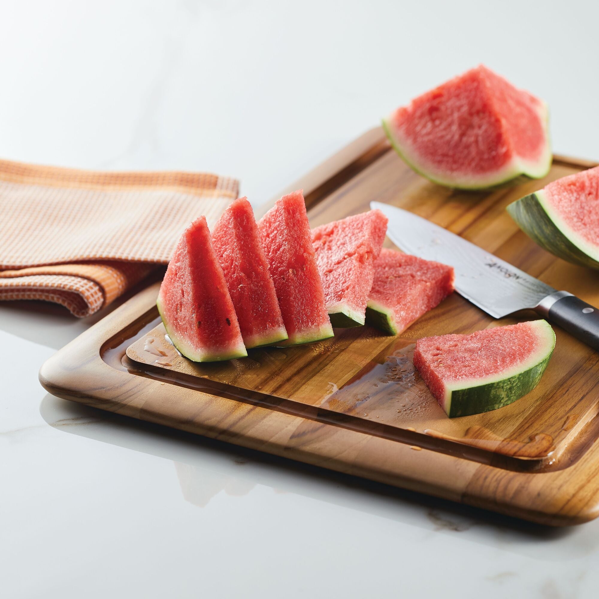Slices of fresh watermelon sit on the Pantryware 20 x 14-Inch Reversible Teak Cutting Board with a knife. An orange and white towel is folded on the white countertop nearby.