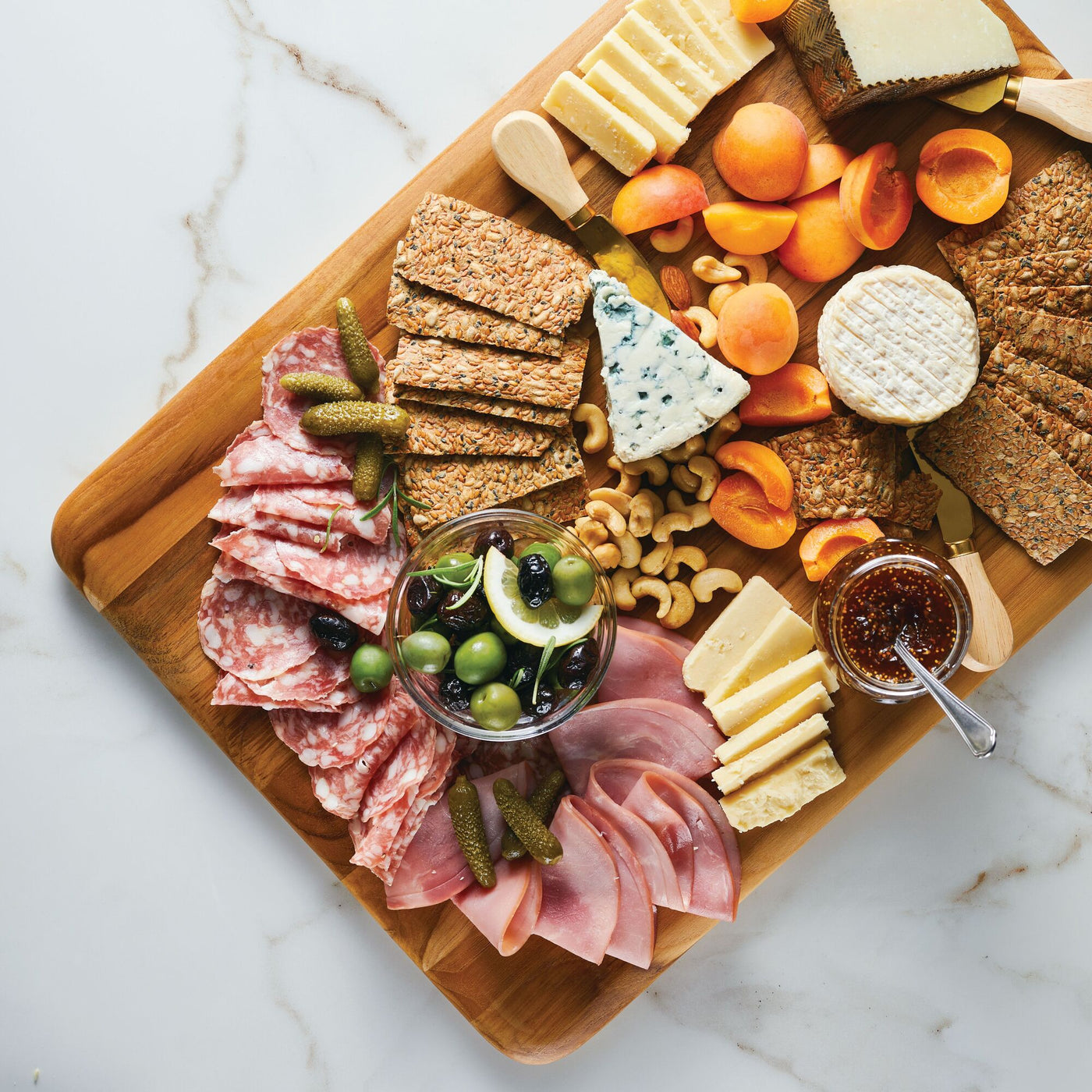 The Pantryware 20 x 14-Inch Reversible Teak Cutting Board showcases cheeses, crackers, meats, pickles, olives, apricots, nuts, and a small bowl of jam on a white marble surface.