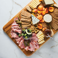 The Pantryware 20 x 14-Inch Reversible Teak Cutting Board showcases cheeses, crackers, meats, pickles, olives, apricots, nuts, and a small bowl of jam on a white marble surface.