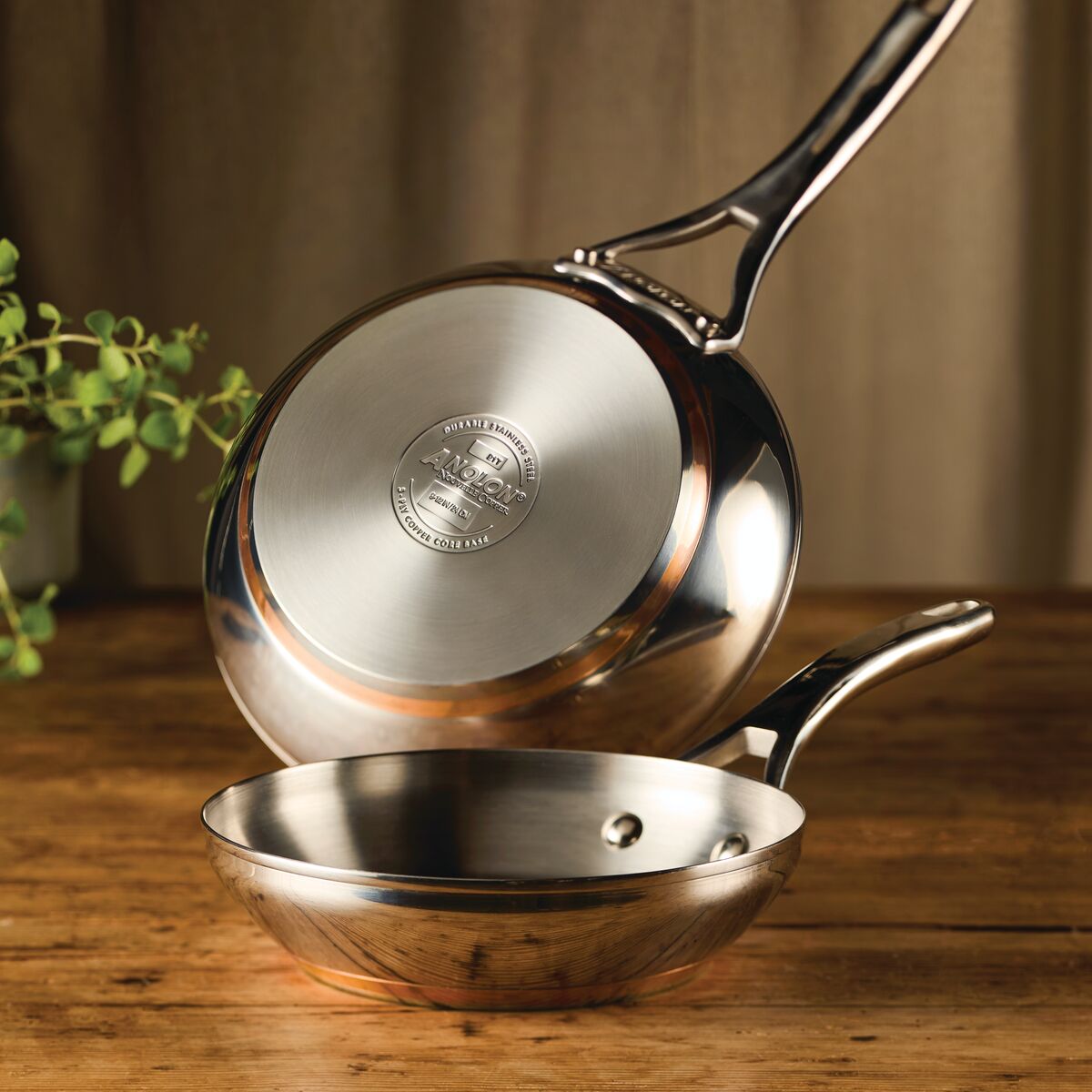 A lifestyle photograph of the Anolon Nouvelle Copper Stainless Steel twin pack frying pan set. One pan is tilted to showcase the polished stainless steel exterior and the thick, five-layer base with a visible copper mid-layer and the Anolon brand etched into the bottom. The second pan sits upright on a rustic wooden surface, highlighting the gleaming stainless steel interior.
