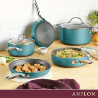A collection of teal cookware is displayed on a kitchen countertop, including pans with sautéing scallops and green beans. A maroon cloth is draped nearby. The background features a white tile backsplash and ingredients like green onions and lemons.