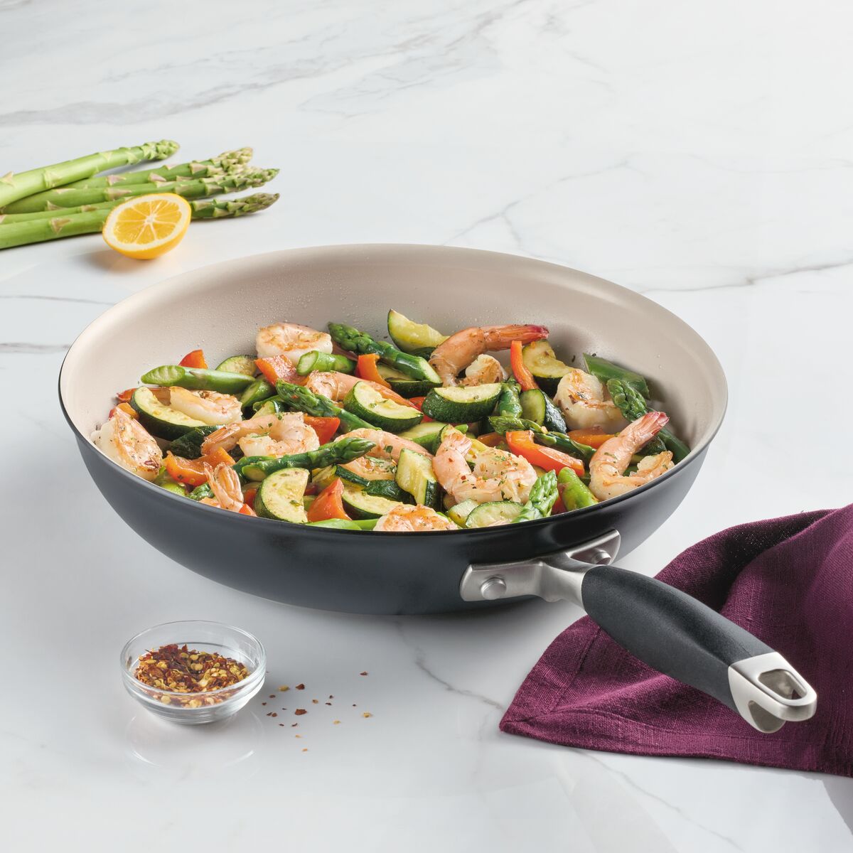 A 12-inch Ceramic Nonstick Frying Pan from Advanced Ceramic holds shrimp, zucchini, carrots, and asparagus on a marble surface. Nearby are red pepper flakes in a bowl, lemon slice, and asparagus spears. A purple napkin highlights the pans elegance beautifully.