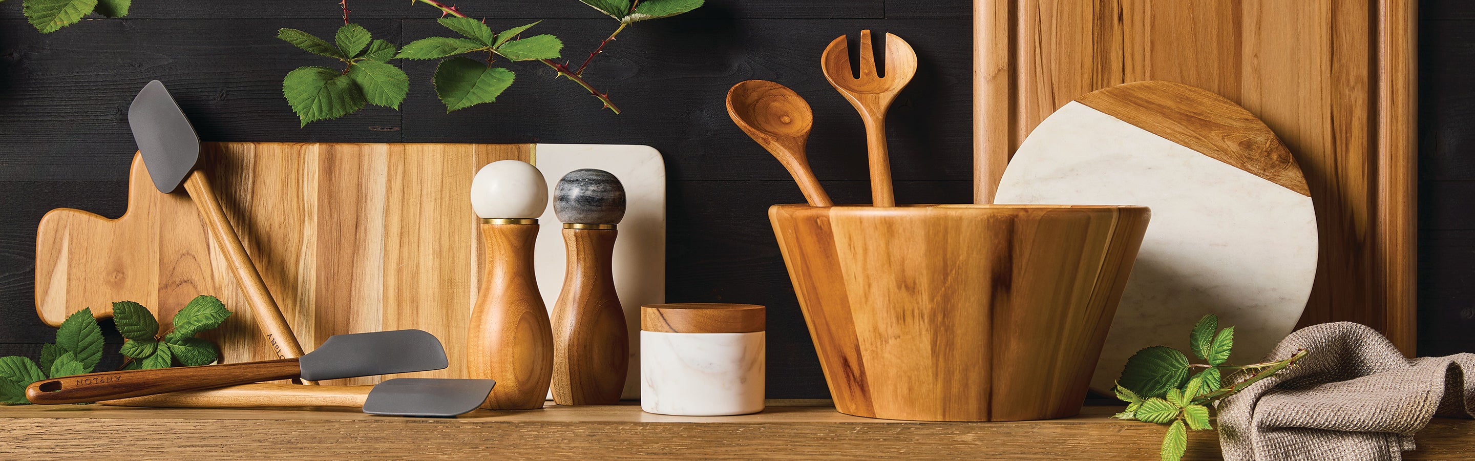 A curated selection of Anolon Pantryware and tools are displayed on a light wooden shelf against a dark background. The collection features a large wooden salad bowl with matching servers, a marble and wood salt cellar, and elegant wooden salt and pepper grinders. To the left, several gray silicone spatulas with wooden handles rest against various wooden and marble cutting boards.