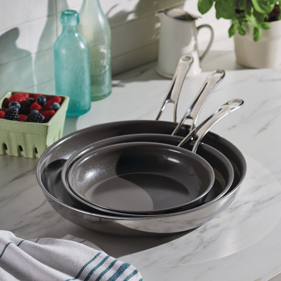 An EverLast Ceramic Stainless Steel 3-Piece Nonstick Frying Pan Set, each with a stainless steel handle, is stacked on a marble countertop. Nearby are a leafy jar, white jug, vintage glass bottles, berry basket, and a blue-striped white towel in the corner.