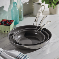 An EverLast Ceramic Stainless Steel 3-Piece Nonstick Frying Pan Set, each with a stainless steel handle, is stacked on a marble countertop. Nearby are a leafy jar, white jug, vintage glass bottles, berry basket, and a blue-striped white towel in the corner.