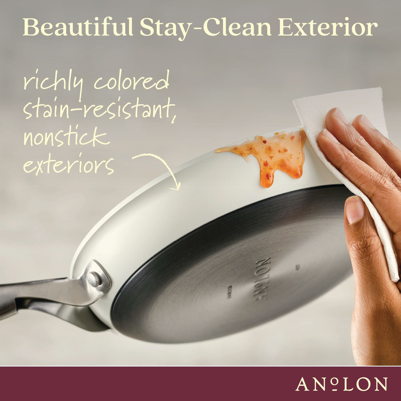 A hand wiping a clean frying pan with a paper towel. The pan has a white, stain-resistant nonstick exterior with a small spill of orange sauce. Text reads, Beautiful Stay-Clean Exterior and richly colored stain-resistant, nonstick exteriors.