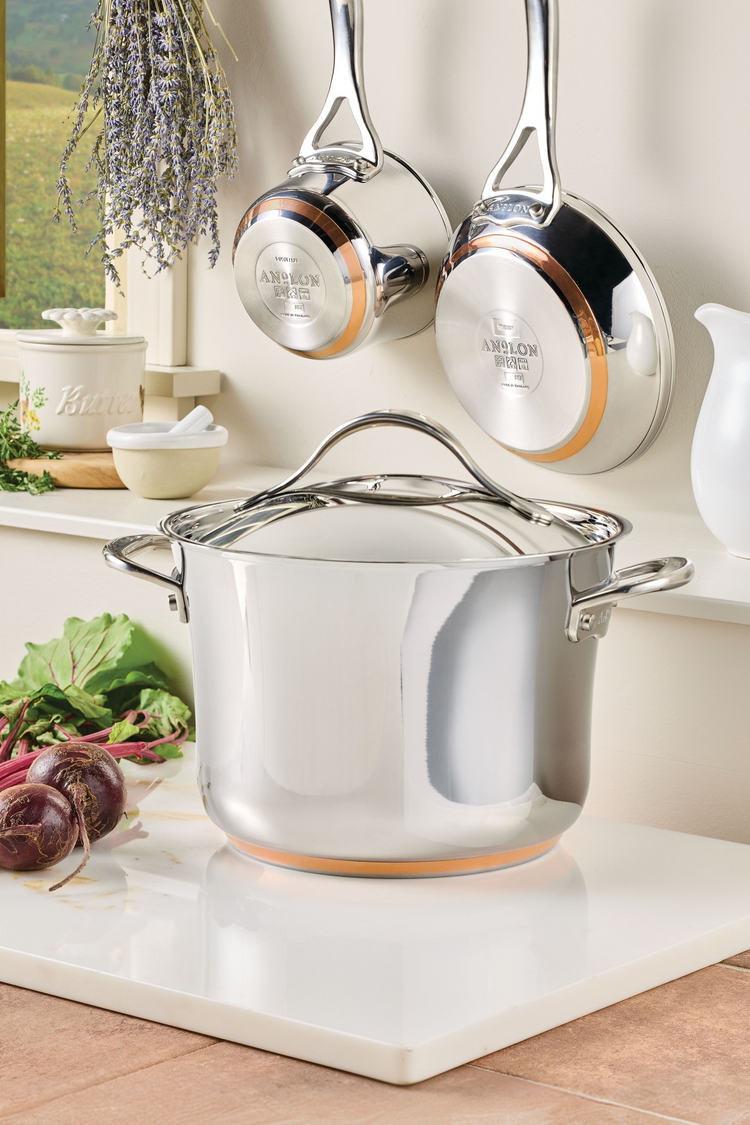 An Anolon Nouvelle Copper stainless steel stockpot with a lid sits on a white marble slab, with a saucepan and frying pan hanging on the wall behind it, featuring visible copper rings on their bases and a bundle of dried lavender in the background.